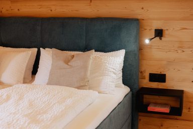 Bett mit blauer Kopfstütze, weißen Kissen und Holzwand im Hintergrund.
