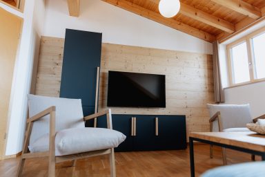 Moderne Wohnzimmereinrichtung mit Fernseher, Holzverkleidung und gemütlichen Stühlen.